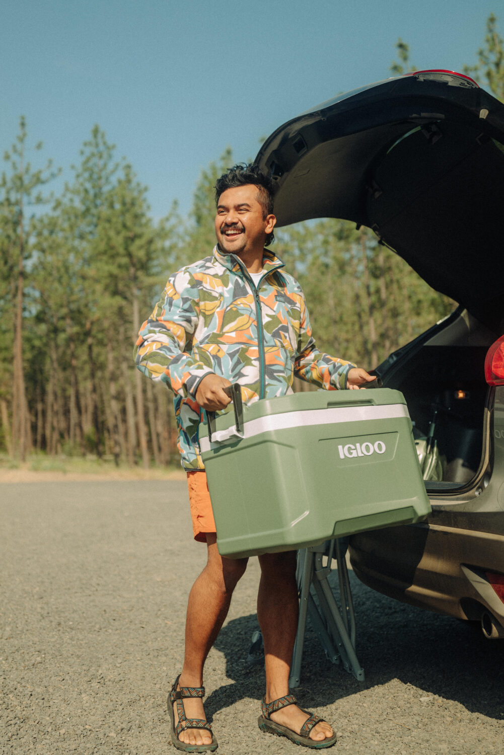 Berty Mandagie holding a green igloo cooler, pulling it from the trunk of a car. He is smiling and ready for a day at the beach