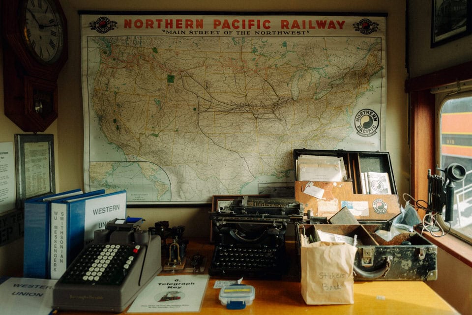 Train memorabilia inside a train car at the Inland NW Train Museum. Poster on teh wall says "Northern Pacific Railway" and there is a typewriting, old suitcase, and phone on the desk.